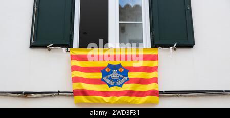 Maó, Menorca, Balearen, Spanien. Farbenfrohe menorquinische Flagge, die vom offenen Fenster eines Altstadthauses herabhängt. Stockfoto
