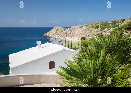 Cala Morell, Menorca, Balearen, Spanien. Blick auf die felsige Landzunge vom Hügel, Dach eines typischen weißen Hauses im Vordergrund. Stockfoto