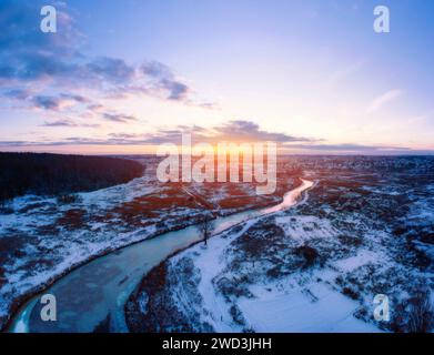 Winterlandschaft mit verwinkelten gefrorenen Flüssen im Feld bedeckt mit Schnee und wunderschönem Sonnenunterganghimmel. Panoramablick aus der Vogelperspektive. Stockfoto