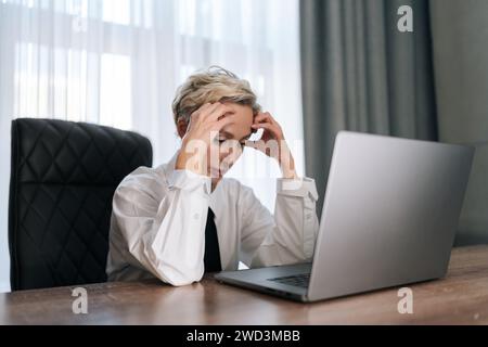 Erschöpfte, verärgerte Geschäftsfrau mittleren Alters, die mit Laptop am Tisch sitzt, Kopf auf Hand lehnt, an Kopfschmerzen leidet, sich depressiv fühlt, unglücklich Stockfoto