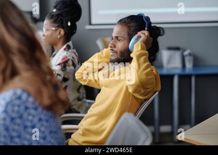 Indischer Büroangestellter sitzt mit Kopfhörern am Schreibtisch, seine weiblichen Kollegen arbeiten am Hintergrund Stockfoto