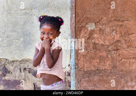 Glückliches afrikanisches Mädchen aus dem Dorf mit Zöpfen und einem großen, zahnlosen Lächeln im Garten an einem Sommertag Stockfoto