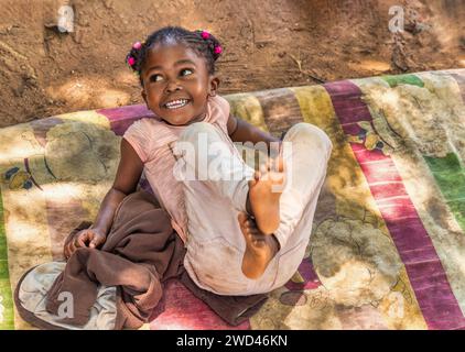 Glückliches afrikanisches Mädchen aus dem Dorf mit Zöpfen und einem großen, zahnlosen Lächeln, das an einem Sommertag auf einer Decke im Hof spielt Stockfoto