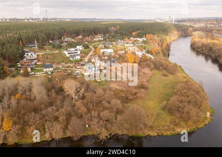 Drohnenfotografie von kleinen Häusern in einem Wald in der Nähe eines Flusses und einer Stadt im Hintergrund während des Herbstvormittags Stockfoto