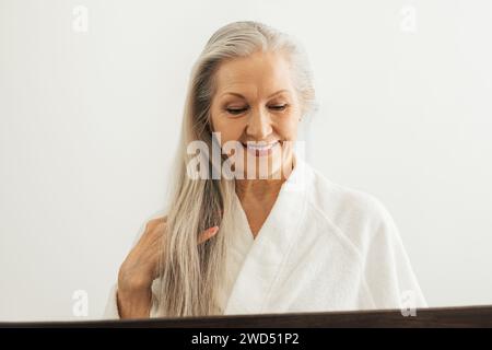 Ältere Frau im Bademantel, die nach unten blickt, während sie ihr langes weißes Haar vor einem Spiegel anpasst Stockfoto