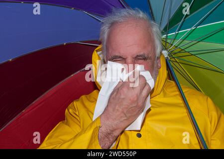 Ein älterer Mann im gelben Regenmantel mit einem bunten Regenschirm trotzt dem schlechten Wetter, obwohl er von einer laufenden Nase geplagt wird. Stockfoto