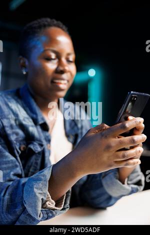 Schwarze Frau sitzt am Tisch und schreibt positiv Nachrichten auf ihrem Handy. Die afroamerikanische Dame lächelt, während sie auf ihrem Mobilgerät im Internet surft und zu Hause moderne Technologie nutzt. Stockfoto