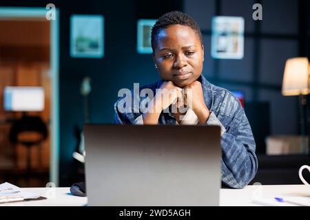 Schwarze Frau, die sich aufgebracht und müde fühlt, schaut auf ihren PC, der auf dem Tisch steht. Durchdachte afroamerikanische Studentin, die Online-Zeitungen mit einem digitalen Laptop liest. Stockfoto
