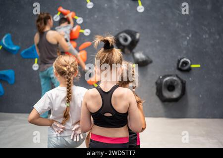 Rückansicht von drei Mädchen in Activewear, die Coach während des Wandkletterkurses beobachten. Sportliche weibliche Kinder beim Klettern. Junge Mädchen repräsentieren Gesundheits- und Fitnesskonzepte. Stockfoto