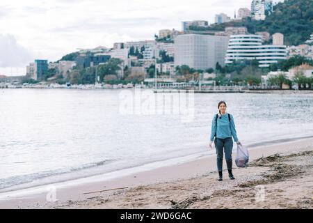 Eine junge Frau mit einer Tüte Müll in Handschuhen steht an einem Sandstrand in der Nähe des Meeres Stockfoto