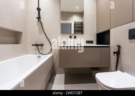 Badezimmer in modernem Stil mit grauen und weißen Fliesen. Es gibt einen großen Spiegel mit leuchtenden Lampen, Tischplatte mit Holzschubladen und Waschbecken, Bad mit Stockfoto