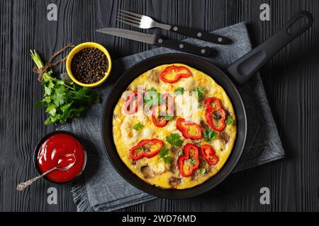 Omelette mit Blumenkohl, rotem Pfeffer, Auberginen und Zuckermais in der Pfanne auf schwarzem Holztisch mit Besteck, Ketchup, Pfeffer und frischem Koriander Stockfoto