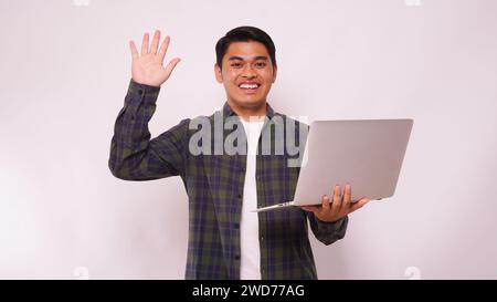 Asiatischer Schüler winkt die Hand, während er den Laptop vor weißem Hintergrund hält Stockfoto