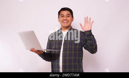 Asiatischer Schüler winkt die Hand, während er den Laptop vor weißem Hintergrund hält Stockfoto