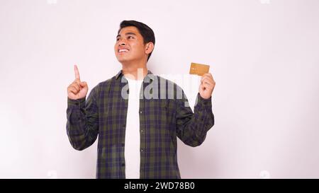 Junger asiatischer Mann lächelt zuversichtlich, während er auf leere Kreditkarte zeigt, die er über grauem Hintergrund hält Stockfoto