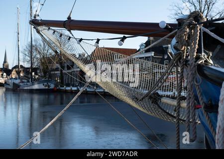 Schiffsbug mit Auslegerausleger und fest geknotetem Auslegernetz vor dem Vormast eines vertäuten Segelschiffes. Eine dünne Eisschicht auf dem Wasser Stockfoto