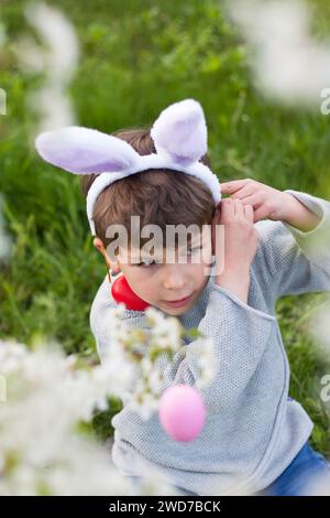 Ostereierjagd. Vorschuljunge, der Hasenohren trägt, sammelt bunte Eier auf der Ostereierjagd im blühenden Garten im Frühjahr. Ostertradition Stockfoto