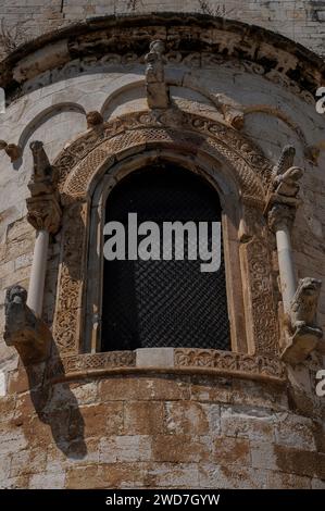 Rundbogenfenster in der zentralen Apsis am östlichen Ende der Kirche der Templer oder Allerheiligen (Chiesa di Ognissanti già dei Templari) in Trani in Apulien, Italien. Die Skulptur umfasst Knüpfarbeiten oder Verflechtungen und die Darstellung von mythischen Kreaturen. Die Kirche wurde in den 1100er Jahren erbaut, wobei die Apsiden und die skulpturalen Verzierungen den 1200er Jahren zugeschrieben wurden Stockfoto