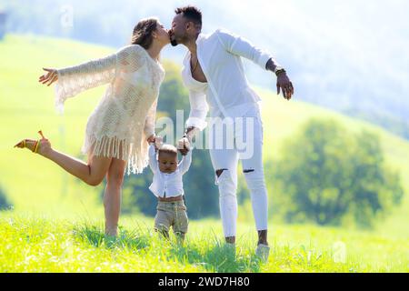 Multiethnische Eltern und gemischtes Baby, das auf Sommergras läuft. Multiethnisches Paar küsst die Natur. Birassisches multiethnisches Baby-Kind erster Schritt Stockfoto