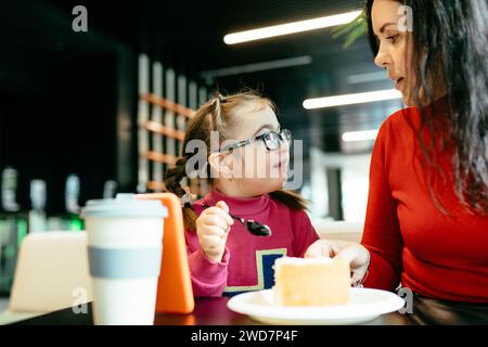 Porträt einer glücklichen Mutter, die sich gegenseitig lächelt, nettes Mädchen mit Down-Syndrom, das Kuchen im Café isst. Happy Family Moments Konzept. Stockfoto