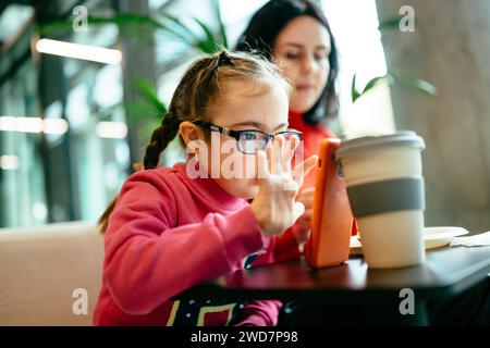 Porträt einer beschäftigten Mutter und eines niedlichen Mädchens mit Down-Syndrom im Café mit Smartphone. Stockfoto