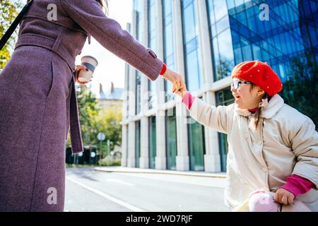 Niedliches kleines Mädchen in Rot trägt Baskenmütze und Mantel in Brille, wirbelt runzelnd und spielt mit ihrer unerkennbaren Mutter in der herbstlichen sonnigen Stadt Stockfoto