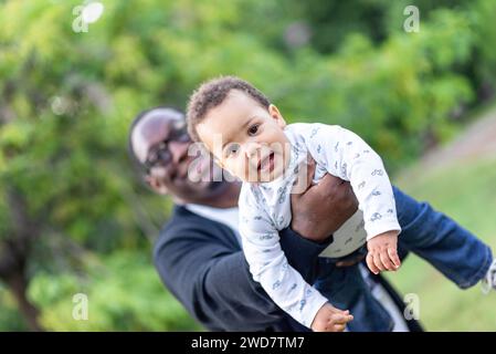 Vater kuschelt niedlichen birassischen Baby-Jungen, kleiner afroamerikanischer Junge, der die Kamera ansieht, wie er sich draußen mit Dad verbündet Stockfoto