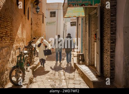 Eine ältere Tunesierin in einem traditionellen Foutah läuft in der alten Medina von Kairouan in Tunesien an zwei Männern und Motorrädern vorbei. Kairouan ist Stockfoto