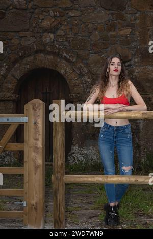 Frau, die sich auf einen neuen Holzzaun vor einem alten Steinhaus mit einer bogenförmigen Holztür lehnt, mit rotem Oberteil und aufgerissener blauer Jeans Stockfoto