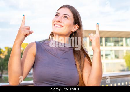 Porträt Junge kaukasische Frau, die mit zwei Daumen nach oben zeigt und draußen lächelnd aussieht. Stockfoto