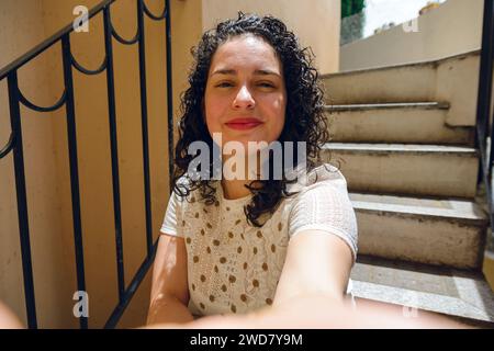 Porträt einer jungen lateinamerikanischen venezolanischen Frau mit Lockenwicklern, Make-up und eleganter Kleidung, die lächelnd im Hotel ankommt und Selfie mit ihrem Handy auf St. macht Stockfoto