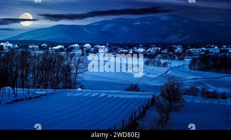 Bergige Landschaft im Winter bei Nacht. Landschaft mit schneebedeckten ländlichen Feldern im Vollmondlicht Stockfoto