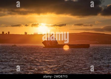Ein Schiff segelt während des goldenen Sonnenuntergangs durch die Gewässer der Stadtküste, mit Sonne am Horizont und einem Sonnenstrahl, der durch die Wolke bricht. Stockfoto