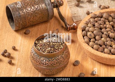 Eine Nahaufnahme einer traditionellen schwarzen Pfeffermühle, gefüllt mit ganzen Pfefferkörnern, auf einem strukturierten Holzhintergrund. Die warme Beleuchtung verbessert die r Stockfoto
