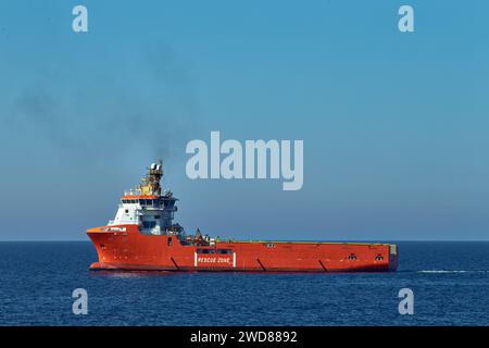 Versorgungsschiff mit roter Plattform an einem hellen Tag am Horizont im blauen Meer, mit Rauch aus dem Abgastrichter und klarem Himmel. Stockfoto