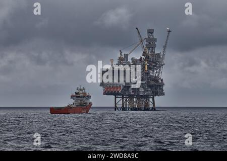 Offshore-Erdöl- und Erdgasplattform mit Versorgungsschiff, das Frachtvorgänge im Meer durchführt, mit dramatischer grauer Meereslandschaft. Stockfoto