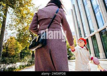 Das kleine Mädchen in Rot trägt Baskenmütze und Mantel in Brille, wirbelt herum und spielt mit ihrer unerkennbaren Mutter auf der sonnigen Straße des Herbsttags Stockfoto