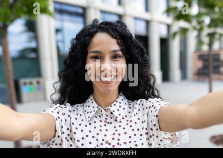 Eine strahlende junge lateinamerikanische Geschäftsfrau, die ein Selfie mit freudigem Ausdruck vor einem modernen Bürogebäude in der Stadt macht. Stockfoto