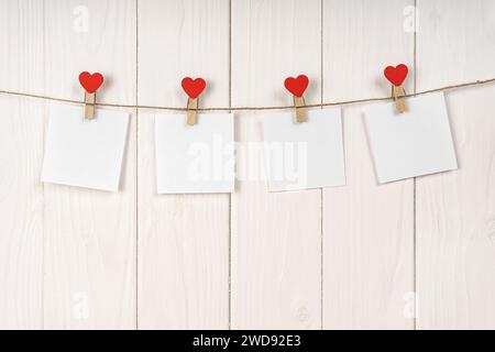 Kleiderschalen mit roten Herzen und leeren Blättern Papier auf Schnur vor weißem hölzernem Hintergrund. Valentinstag, romantisches Konzept. Modell, tem Stockfoto