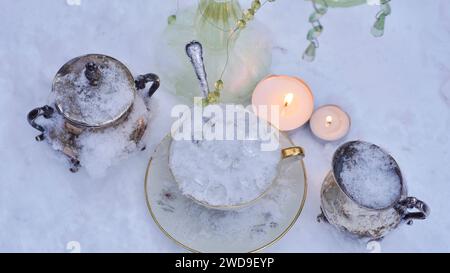 Eine Tasse voller Eis im Schnee. Glaskristalle. Beleuchtete Teelampen. Eine silberne Milch- und Zuckerdose. Tisch für die Eiskönigin. Stockfoto