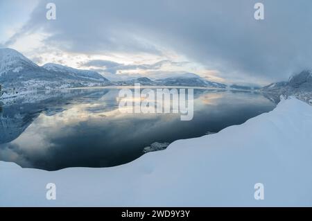 Das Bild fängt eine ruhige Winterszene mit einem glatten, spiegelähnlichen Fjord ein, der die sanften Farben des Abendhimmels in schneebedeckter Umgebung reflektiert. Stockfoto