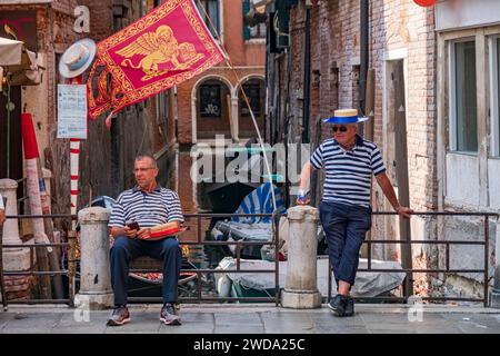 03.07.2018, Venedig, Italien. Alte und junge Gondoliere in traditioneller Kleidung stehen auf dem Pier mit venezianischer Flagge und Kanälen im Hintergrund Stockfoto