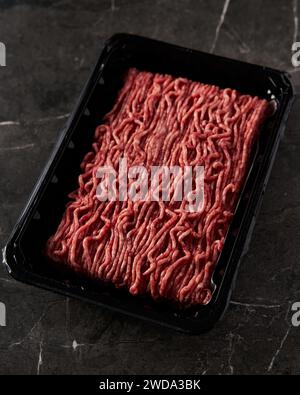 Rohes und frisches Hackfleisch in einem schwarzen Plastikbehälter aus dem Laden, geöffnet. Auf dunklem Marmorhintergrund. Minimalistisches Essensfoto, dezent. Stockfoto
