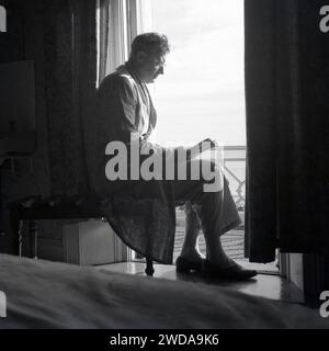 1960er Jahre, historisch, in einem Hotelzimmer, am frühen Morgen und ein Gentleman in Schlafanzug und einem leichten Baumwollmantel, der ein Buch auf dem Balkon mit Blick auf das Meer liest, England, Großbritannien. Stockfoto