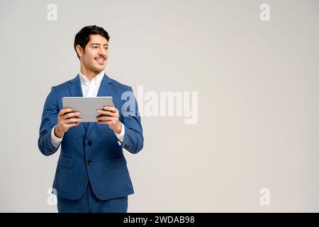 Der hübsche hispanische Mann im blauen formalen Anzug lächelt während er das Tablet benutzt, bietet ein ansprechendes Bild der Verwendung von Technologie im täglichen Geschäft, das Konzept der Integration von Technologie in das Berufsleben Stockfoto