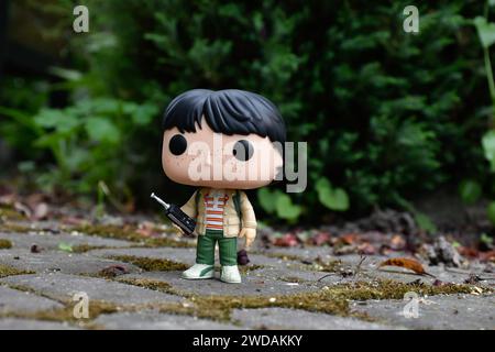 Funko Pop Actionfigur von Mike mit Walkie-Talkie aus der beliebten Horror-TV-Serie Stranger Things. Grüne Blätter, moosige Steinstraße, Frühlingsgarten. Stockfoto