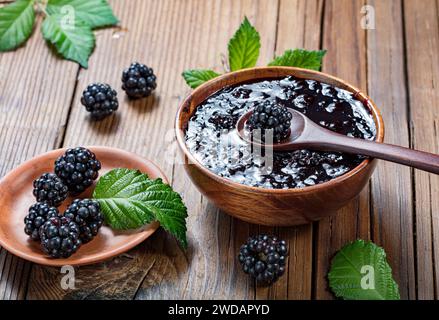 brombeermarmelade in Holzschüssel und Beeren und Brombeerblätter auf hölzernem Hintergrund Stockfoto