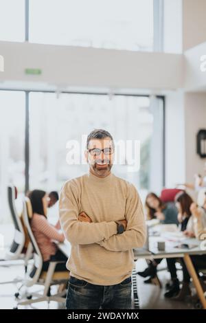 Porträt eines fröhlichen Geschäftsmannes mittleren Alters, der selbstbewusst in einem hellen, modernen Bürobereich steht und sein Team hinter ihm arbeitet. Stockfoto