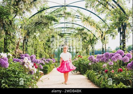 Entzückendes kleines Mädchen, das an einem schönen sonnigen Tag in einem wunderschönen Blumengarten spielt, mit weißer Bluse und einem knallrosa Tutu-Rock. Gasse mit Riesenalliu Stockfoto