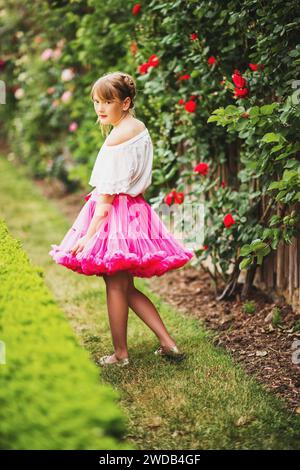 Außenporträt des entzückenden kleinen 9-10-jährigen Mädchens mit weißem Hemd und rosa Tutu-Rock, das im wunderschönen Sommergarten spielt Stockfoto